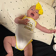 Haven joined the competition — help win amazing prizes! baby, infant, smiling, yellow_bow, onesie, text_on_clothing, pillow, patterned_pillow, lying_down, happy, cute, child, indoor, skin, headband, reaching, open_mouth, cheerful, black_surface, yellow_trim