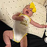 baby, infant, smiling, yellow_bow, onesie, text_on_clothing, pillow, patterned_pillow, lying_down, happy, cute, child, indoor, skin, headband, reaching, open_mouth, cheerful, black_surface, yellow_trim