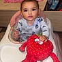 baby, high_chair, plush_toy, strawberry, smiling_toy, indoor, blanket, vase, decor, child, person, clothing, furniture, face, head, hands, wood_floor, room, soft_toy, cute