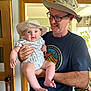 Henry is registered to the contest to win money with this photo: baby, man, hat, glasses, smile, indoor, holding, onesie, patterned_clothing, mirror, door, plants, toy, person, face, happy, casual_clothing, home, child, adult