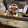 baby, cap, chair, child, container, curious, floor, furniture, green_shirt, holding, home, indoor, person, plastic_balls, playpen, standing, stuffed_animal, table, toddler, toy