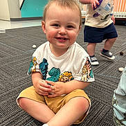 Benjamin is registered to the contest to win money with this photo: toddler, child, smiling, sitting, indoor, carpet, toy, shorts, tshirt, play, daycare, happy, person, floor, cute, young_child, kids, background, expression, casual_clothing