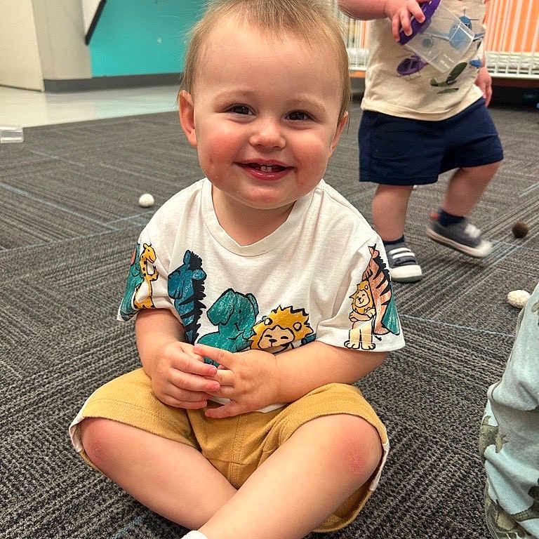 Benjamin is registered to the contest to win money with this photo: toddler, child, smiling, sitting, indoor, carpet, toy, shorts, tshirt, play, daycare, happy, person, floor, cute, young_child, kids, background, expression, casual_clothing