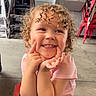 child, toddler, curly_hair, smiling, indoor, casual_clothing, pink_shirt, blue_jeans, happy, posing, fingers, cheeky, stacked_chairs, concrete_floor, signage, red_chairs, playful, portrait, cute, young_child
