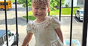Iris is registered to the contest to win money with this photo: child, toddler, curly_hair, smiling, balcony, outdoor, daylight, green_tree, parking_lot, car, school_bus, white_shoes, shorts, top, playful, happy, person, summer_clothing, railing, sidewalk