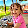 child, toddler, curly_hair, pink_shirt, smiling, outdoor, picnic_table, water_bottle, juice_pouch, covered_pavilion, greenery, sky, wood, daylight, happy, person, casual_clothing, park, nature, portrait