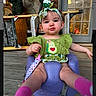 baby, baby_clothing, candid, casual, child, curious_expression, cute, door, green_outfit, headband, headwear, home, infant, infant_toys, outdoor, person, pink_socks, purple_seat, seated, wooden_porch