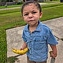 Mason is registered to the contest to win money with this photo: toddler, child, banana, barefoot, denim_shirt, shorts, sidewalk, grass, suburban, residential_area, toy, serious_expression, outdoor, person, greenery, house, curb, summer, casual_clothing, young_child