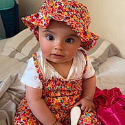 Elyna a rejoint le concours — aidez-le/la à gagner de superbes lots ! baby, bed, blanket, child, chubby_cheeks, colorful_clothing, curious_expression, cute, floral_hat, foot, hairbrush, hat, indoors, infant, person, playful, portrait, sitting, smiling, soft_lighting