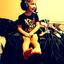 Jasmine joined the competition — help win amazing prizes! child, headset, bed, black_shirt, text_on_clothing, smiling, sitting, legs, barefoot, indoor, window, blinds, clutter, toys, controller, person, young_child, hair_up, happy, casual