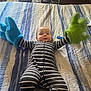 Alakai joined the competition — help win amazing prizes! baby, child, striped_onesie, plush_gloves, blue_glove, green_glove, bed, bedspread, striped_pattern, indoor, infant, person, cute, smiling, playful, furniture, cozy, reaching, soft_texture, home