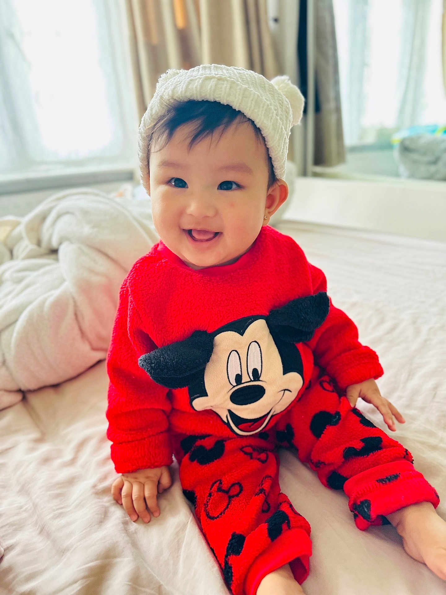 Ngaugin is registered to the contest to win money with this photo: baby, bed, child, cozy, cute, happy, hat, indoor, infant, knitted_hat, mickey_mouse, person, playful, portrait, red_clothing, sitting, smiling, toddler, warm_clothing, young_child