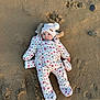 baby, infant, child, sand, beach, winter_clothing, snowsuit, beanie, pompom, floral_pattern, mittens, portrait, lying_down, blue_eyes, fur_trim, footprints, stone, outdoor, cozy, cute