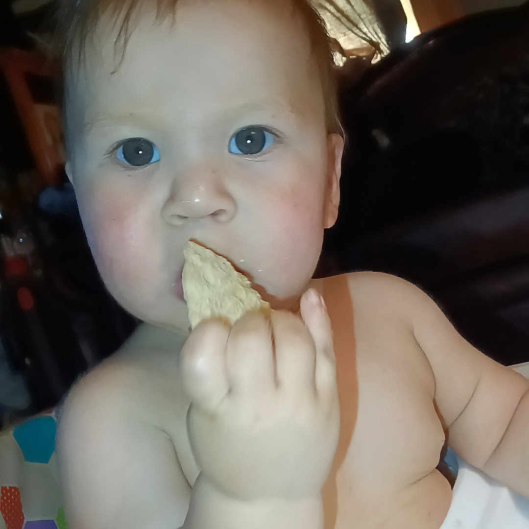 Christian is registered to the contest to win money with this photo: baby, child, infant, portrait, sitting, eating, cracker, snack, hand, blue_eyes, smiling, face, bare_shoulders, indoor, home, couch, blanket, toy, close_up, photograph