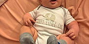 Noah is registered to the contest to win money with this photo: baby, infant, onesie, socks, face, expression, lying_down, bed, sheet, cute, child, skin, hand, footwear, sleepwear, indoors, person, newborn, funny_face, soft_fabric