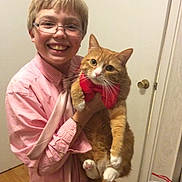 Coco is registered to the contest to win money with this photo: child, boy, glasses, smile, cat, orange_cat, bow_tie, pet, indoor, door, doorknob, hardwood_floor, plaid_shirt, holding, whiskers, paws, portrait, happy, home, camera