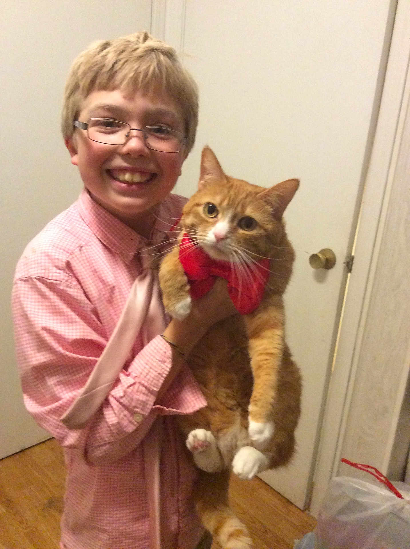 Coco is registered to the contest to win money with this photo: child, boy, glasses, smile, cat, orange_cat, bow_tie, pet, indoor, door, doorknob, hardwood_floor, plaid_shirt, holding, whiskers, paws, portrait, happy, home, camera