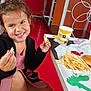 barefoot, black_jacket, burger, casual, child, cup, fast_food, food, fries, girl, happy, indoor, pink_dress, playful, red_floor, restaurant, seat, smiling, snack, table
