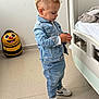 child, denim_jacket, denim_pants, sneakers, nike, hospital_bed, bee_suitcase, floor, standing, side_view, curly_hair, indoor, person, casual_clothing, toy, luggage, white_wall, carpet, focused, young_child