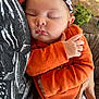 Mac is registered to the contest to win money with this photo: baby, sleeping, infant, orange_clothing, headband, person, hand, closeup, face, peaceful, cozy, cuddling, patterned_shirt, skin, outdoor, natural_light, relaxation, portrait, cute, soft_texture