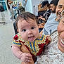baby, infant, person, smiling, holding, clothing, red_dress, indoor, people, seated, waiting_area, luggage, adult, happy, portrait, hand, patterned_clothing, public_space, curly_hair, bright