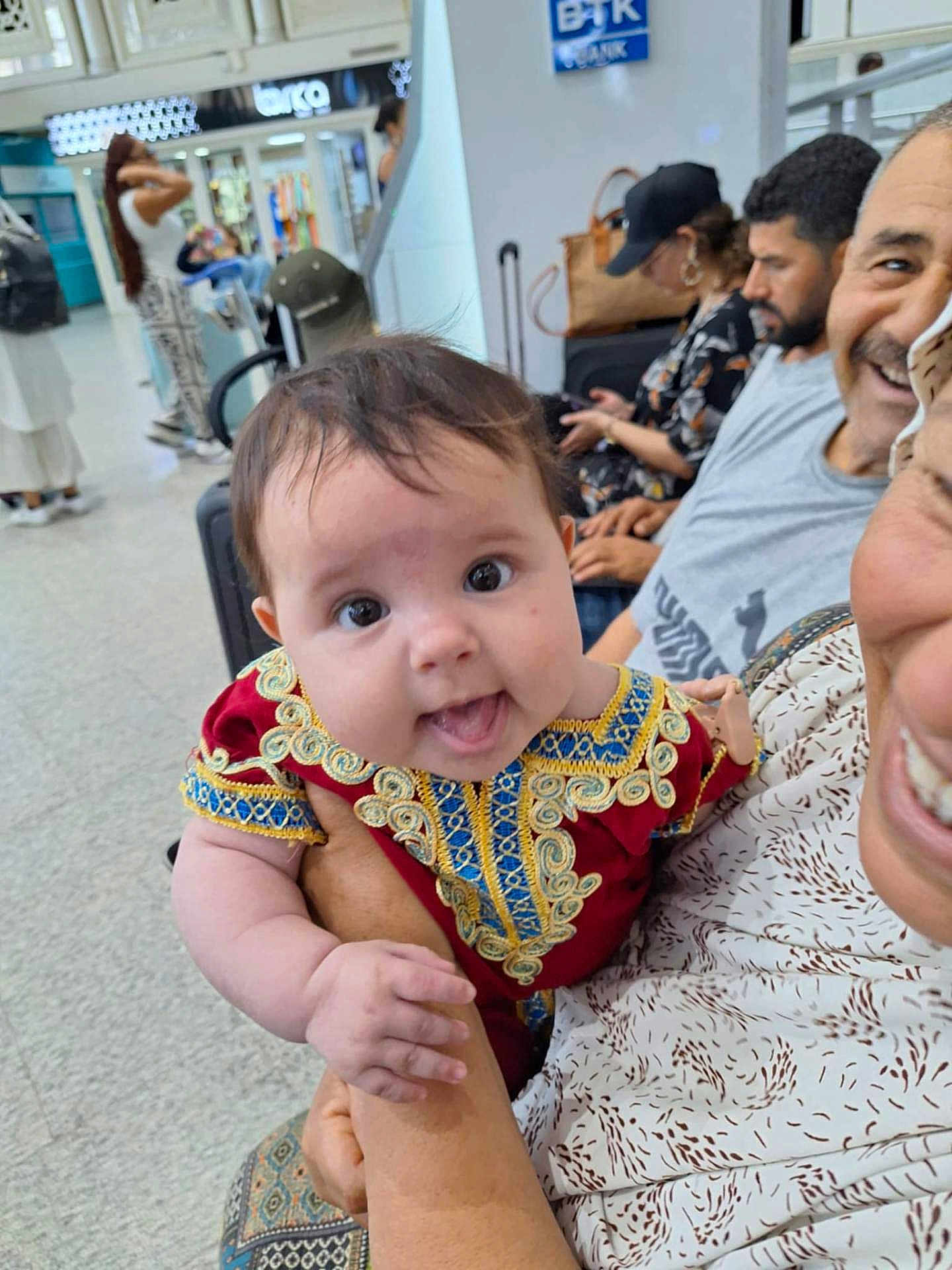 Sarah participe au concours pour gagner de l'argent avec cette photo : baby, infant, person, smiling, holding, clothing, red_dress, indoor, people, seated, waiting_area, luggage, adult, happy, portrait, hand, patterned_clothing, public_space, curly_hair, bright