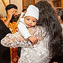 baby, beard, blurred_background, ceremony, child, closeup, event, glasses, happy, hat, holding, indoor, lace_dress, long_hair, man, people, portrait, religious_clothing, smile, woman