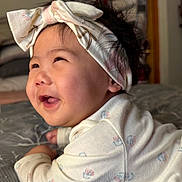Raelynn is registered to the contest to win money with this photo: baby, infant, headband, smiling, happy, cute, lying_down, bed, blanket, pattern, indoors, child, face, cheeks, hair, clothing, soft_light, portrait, closeup, toddler