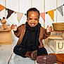 child, toddler, smiling, brown_jacket, black_scarf, boots, floor, wooden_blocks, stuffed_animals, decorations, bunting_flags, hat, basket, indoor, happy, cute, playful, person, portrait, warm_clothing