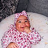 baby, infant, child, smiling, floral_outfit, pink_clothing, headband, bow, thumb_sucking, portrait, face, eyes, cheeks, sitting, cushion, blanket, sofa, indoor, happy, photography