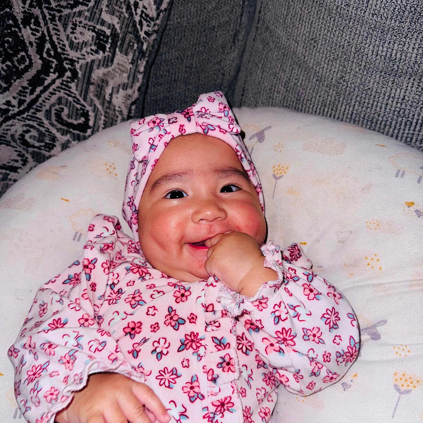 Dayani is registered to the contest to win money with this photo: baby, infant, child, smiling, floral_outfit, pink_clothing, headband, bow, thumb_sucking, portrait, face, eyes, cheeks, sitting, cushion, blanket, sofa, indoor, happy, photography