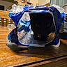 cat, black_cat, plastic_bag, table, wood, indoor, kitchen, pet, animal, curious, container, food_packaging, furniture, domestic, collar, shadow, dark, glowing_eyes, household, playful