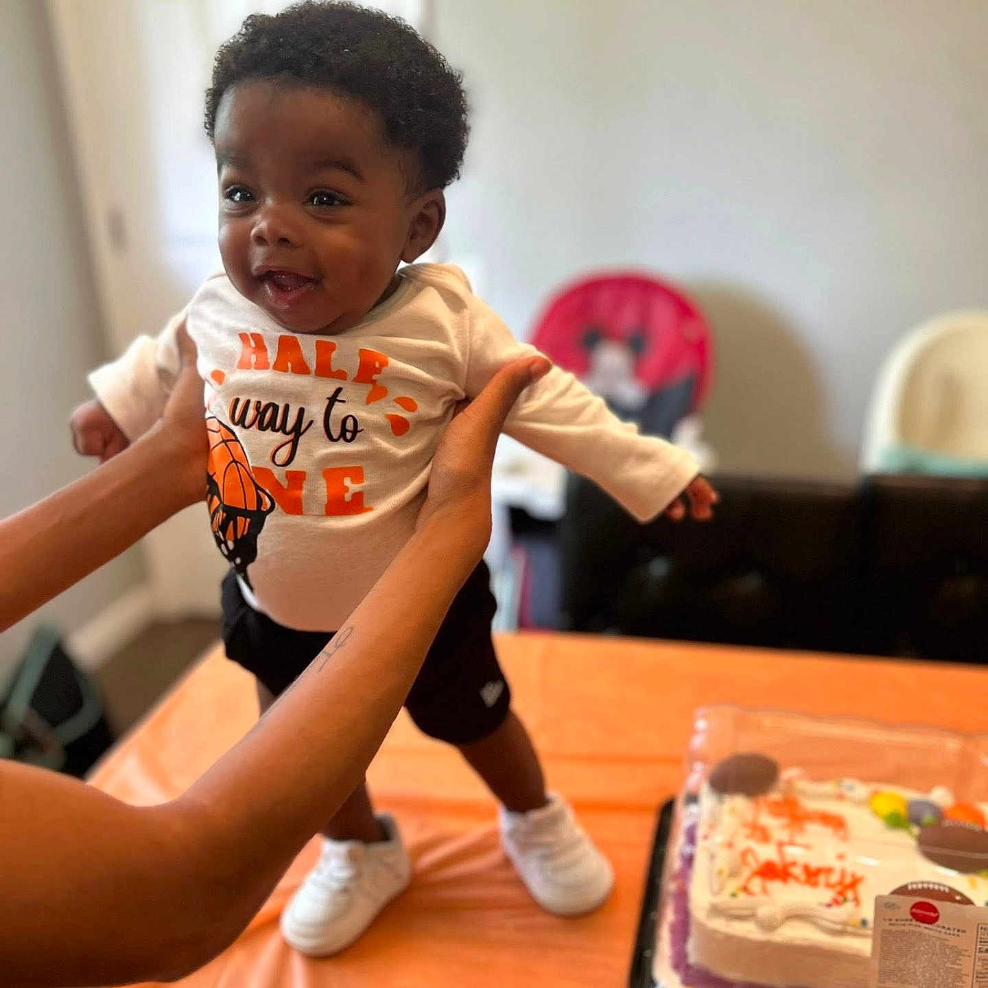 Jakory is registered to the contest to win money with this photo: baby, black_shorts, cake, celebration, child, curly_hair, dessert, food, hands, happy, holding, indoor, infant, orange, party, person, shirt, smiling, sneakers, table