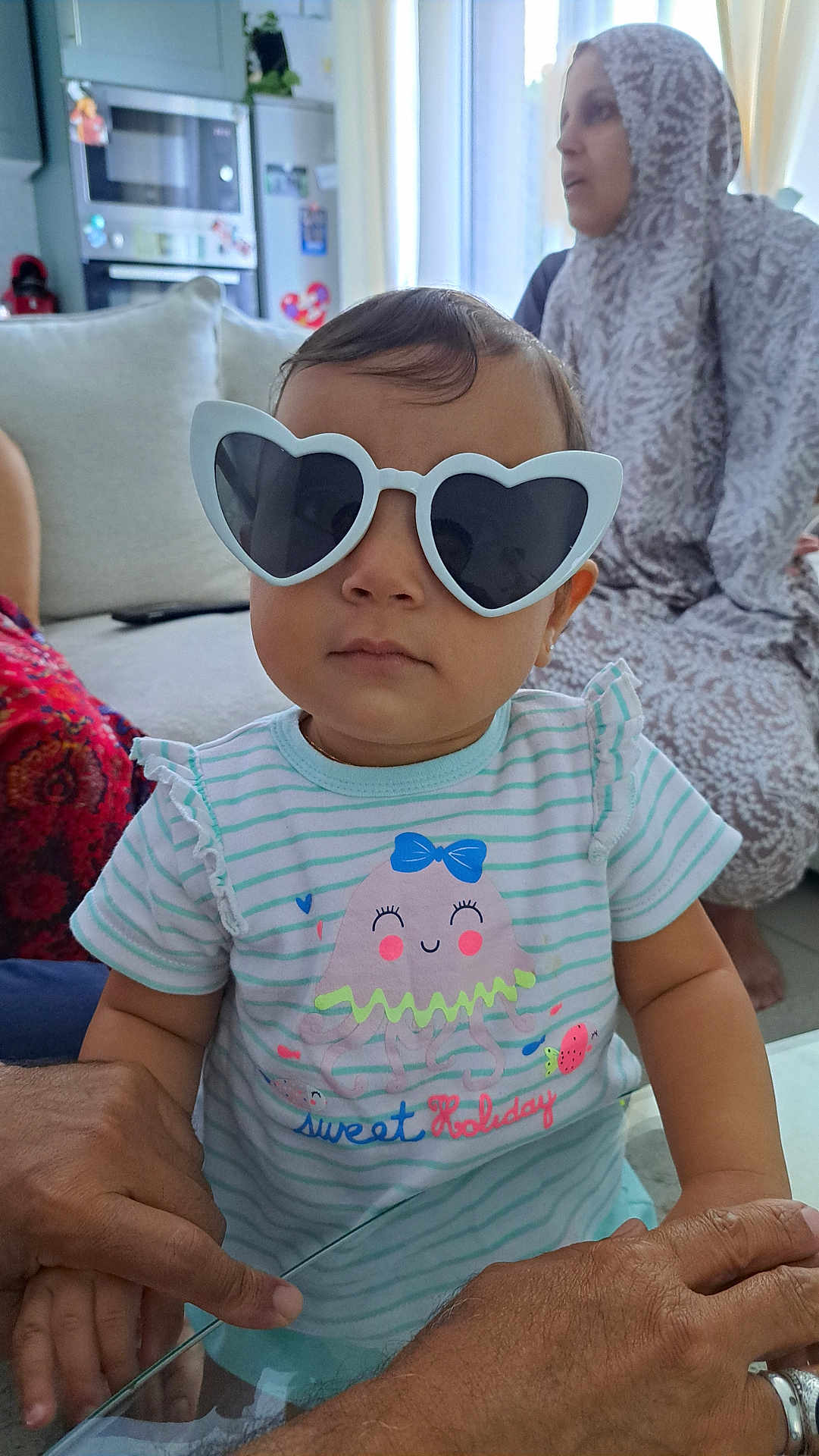 Inaya a rejoint le concours — aidez-le/la à gagner de superbes lots ! toddler, child, glasses, heart_shaped_glasses, striped_shirt, jellyfish_graphic, indoor, living_room, adult_hands, woman, hijab, sofa, couch, table, person, portrait, cute, fashion, casual, family
