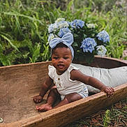 Brianna is registered to the contest to win money with this photo: baby, child, headband, blue_bow, wooden_trough, flowers, hydrangea, greenery, outdoor, nature, grass, plants, cute, sitting, infant, portrait, smiling, white_clothing, pillow, leaf_litter