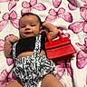 baby, infant, blanket, butterfly_pattern, red_handbag, clothing, black_top, white_shorts, patterned_shorts, person, lying_down, cute, fashion, accessory, soft_texture, indoors, child, portrait, smiling, handbag