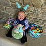 toddler, child, bunny_ears, stuffed_animal, easter_eggs, basket, candy, toy, stone_wall, bench, smiling, cute, indoors, plush, clothing, headband, happy, sitting, holiday, celebration