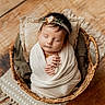 baby, newborn, sleeping, wrapped, blanket, wicker_basket, headband, flower, wooden_floor, rug, texture, peaceful, cute, infant, portrait, indoors, soft_lighting, hands, cozy, resting