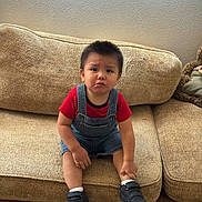 Marbella joined the competition — help win amazing prizes! toddler, child, couch, denim_overalls, red_shirt, black_shoes, wooden_floor, indoor, window_blinds, serious_expression, sitting, person, home_interior, furniture, young_child, short_hair, casual_clothing, floor, living_room, cute