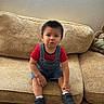 toddler, child, couch, denim_overalls, red_shirt, black_shoes, wooden_floor, indoor, window_blinds, serious_expression, sitting, person, home_interior, furniture, young_child, short_hair, casual_clothing, floor, living_room, cute