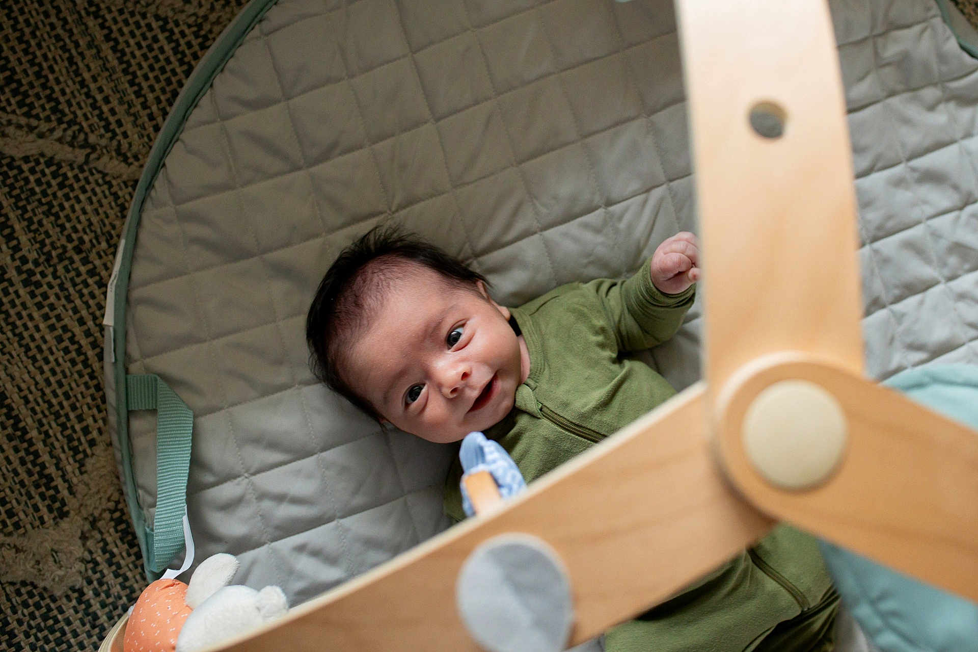 Lashaun is registered to the contest to win money with this photo: baby, infant, green_onesie, quilted_mat, wooden_baby_gym, soft_toy, smiling, face, child, playtime, indoors, happy, cute, lying_down, floor_mat, baby_toys, wooden_toy, close_up, head, person