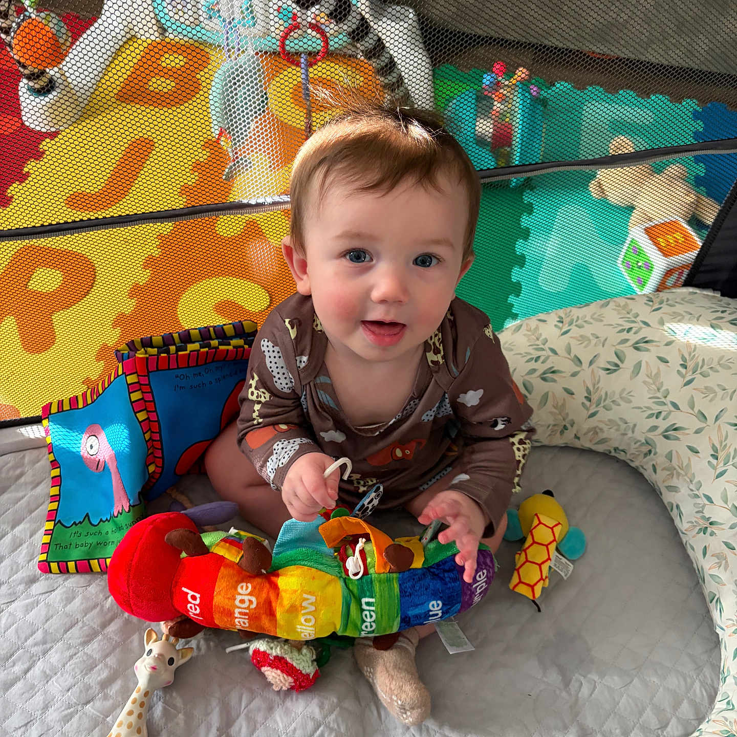 Blaize is registered to the contest to win money with this photo: baby, child, playpen, toy, colorful, plush_toy, giraffe_toy, rainbow, smiling, indoor, playmat, infant, cute, sitting, happy, face, soft_toys, clothing, person, nursery