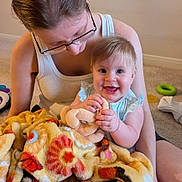 Emma joined the competition — help win amazing prizes! baby, child, woman, glasses, blanket, toy, indoor, carpet, floor, smile, happy, holding, sitting, play, colorful, clothing, person, parent, infant, family