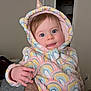 Emma is registered to the contest to win money with this photo: baby, child, unicorn_hoodie, rainbow_pattern, indoor, cute, tongue_out, blue_eyes, hood, soft_clothing, person, infant, hand, facial_expression, warm_clothing, home, door, wall, playful, portrait