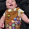 baby, child, smiling, laughing, infant, overalls, badge, brown_clothing, patterned_shirt, cushion, black_background, happy, cute, portrait, indoor, young_child, person, skin, face, expression