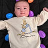 baby, infant, child, plastic_eggs, colorful, black_blanket, cute, smiling, sweater, bunny_illustration, text_on_clothing, lying_down, indoors, soft_lighting, portrait, happy, young_child, playful, clothing, closeup