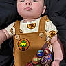 Grayson is registered to the contest to win money with this photo: baby, child, infant, face, eyes, clothing, overall, badge, pattern, brown, skin, arm, cute, portrait, expression, lying_down, black_background, button, fabric, person