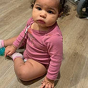 Reason joined the competition — help win amazing prizes! child, toddler, baby, sitting, wooden_floor, pink_onesie, socks, pacifier, stroller, stroller_wheel, curly_hair, topknot, diaper, indoor, portrait, looking_at_camera, serious_expression, hand, flooring, young_child