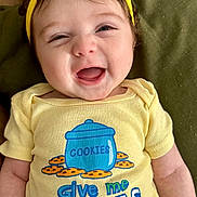 Elodie joined the competition — help win amazing prizes! baby, smiling, yellow_headband, yellow_onesie, cookie_jar_graphic, text_on_clothing, cushion, pillow, indoor, infant, happy, cute, child, portrait, clothing, headwear, lying_down, face, close_up, playful