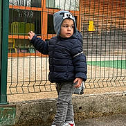 Timothe a rejoint le concours — aidez-le/la à gagner de superbes lots ! child, kid, hat, jacket, fence, outdoor, shoes, sneakers, jeans, playground, building, window, side_view, standing, cold_weather, casual_clothing, curious, young, person, urban
